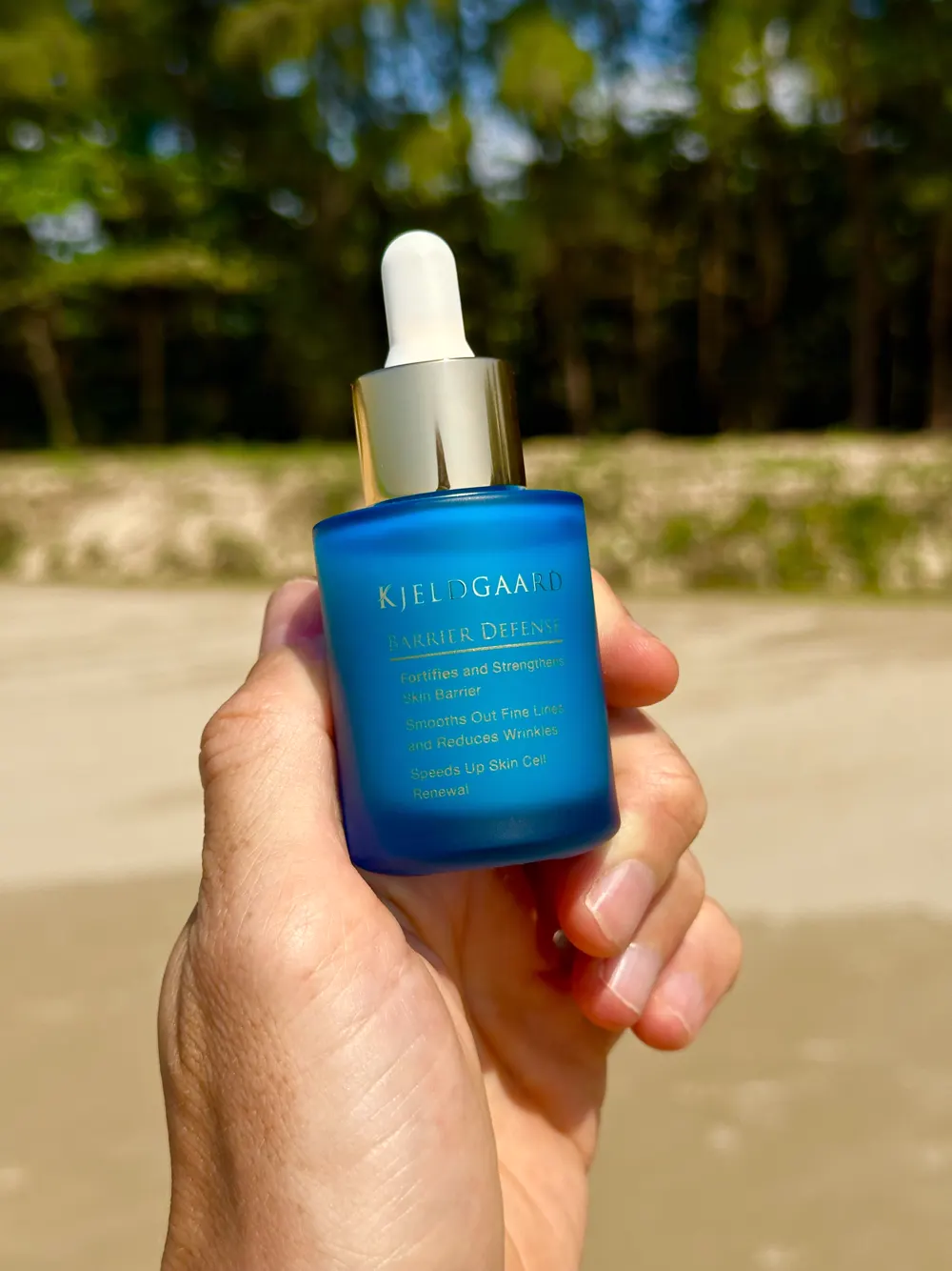 Kjeldgaards Barrier Defense Serum — frosted blå glasflaske med guld-pipette, holdt i hånden udendørs.